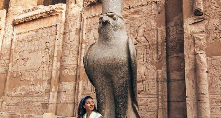 Een persoon die naast een groot stenen standbeeld van een valk staat voor een oude Egyptische muur met hiërogliefen.