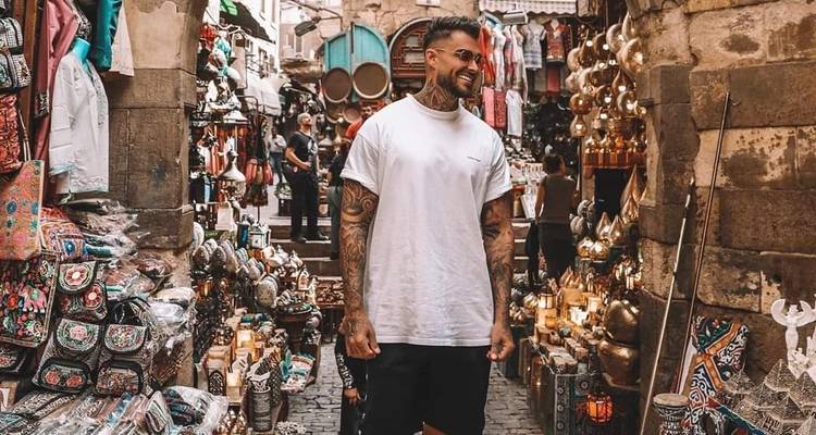 Mann lächelt, während er durch einen belebten Basar mit bunten Gegenständen geht.