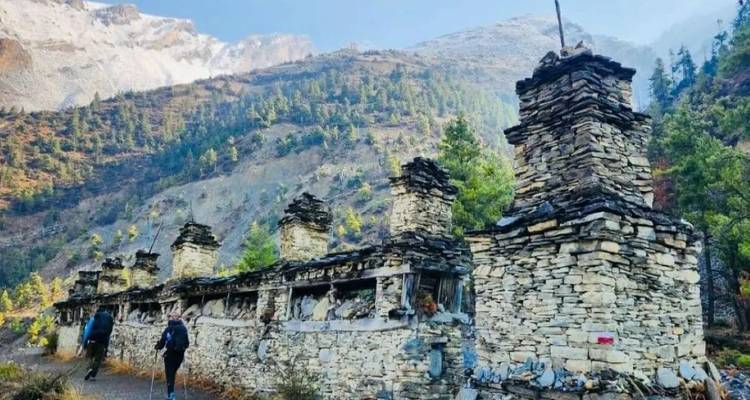 Wanderer gehen an einer langen Reihe von verwitterten steinernen Mani-Mauern in einem bewaldeten Himalaya-Tal vorbei.