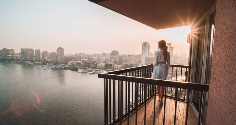 Woman standing on a balcony overlooking a river cityscape at sunrise.