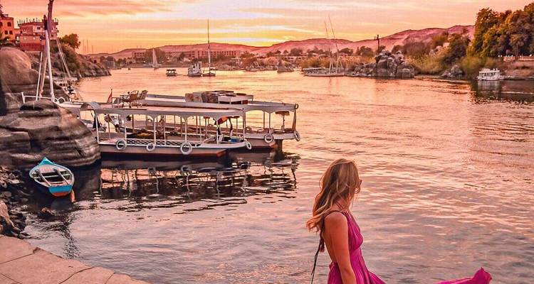 A woman in a pink dress standing by the river with boats and a sunset view.