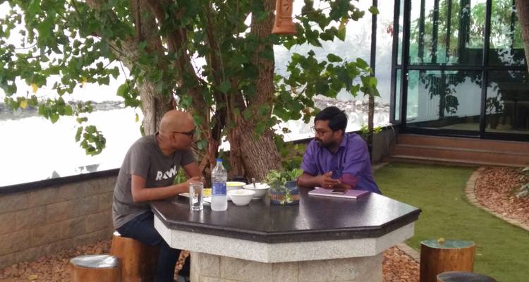 Zwei Personen unterhalten sich an einem Steintisch unter einem Baum.