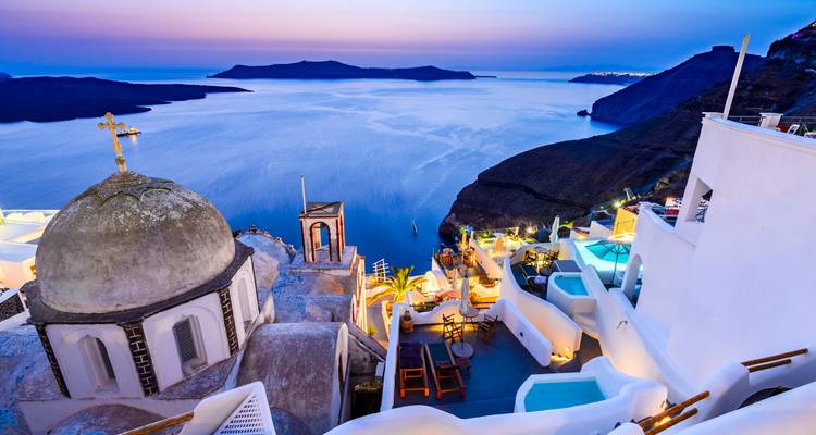 Vista elevada del atardecer sobre los acantilados encalados de Santorini y la caldera con piscinas brillantes y cúpulas.
