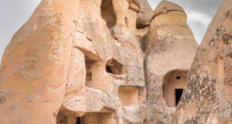 Viviendas de roca desgastada talladas en acantilados volcánicos rosados en Capadocia bajo cielos grises.