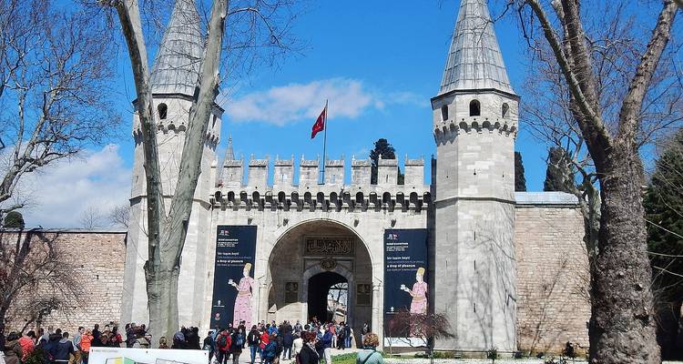 Menschenmengen betreten das steinerne Tor des Topkapi-Palastes unter der türkischen Flagge und Türmen an einem klaren Tag.