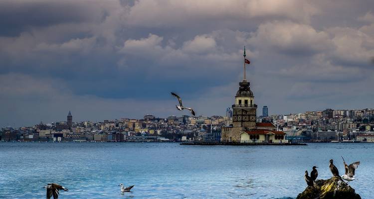 Blick über den Bosporus auf den Jungfernturm mit Stadtskyline und Seevögeln unter dramatischem Himmel.