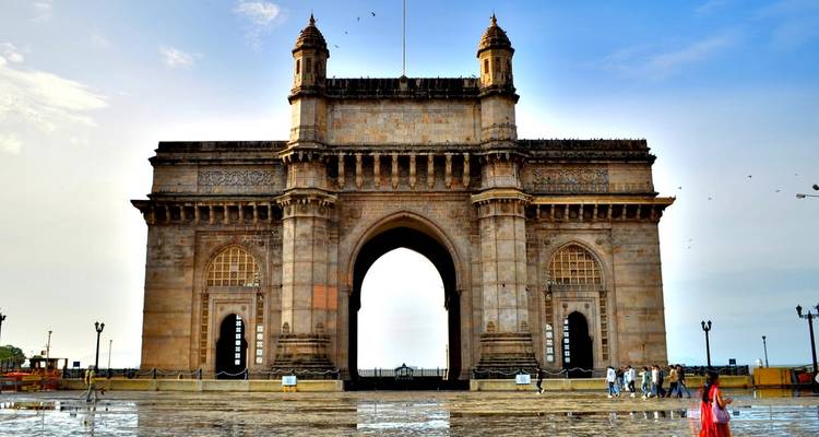 Front view of the Gateway of India monument with a few visitors walking across a wet plaza under a clear sky