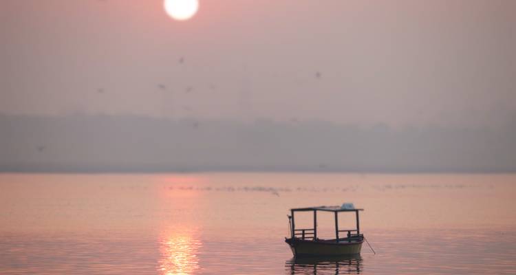 Solitary wooden boat floating on a misty river at sunrise with soft pink sun overhead.