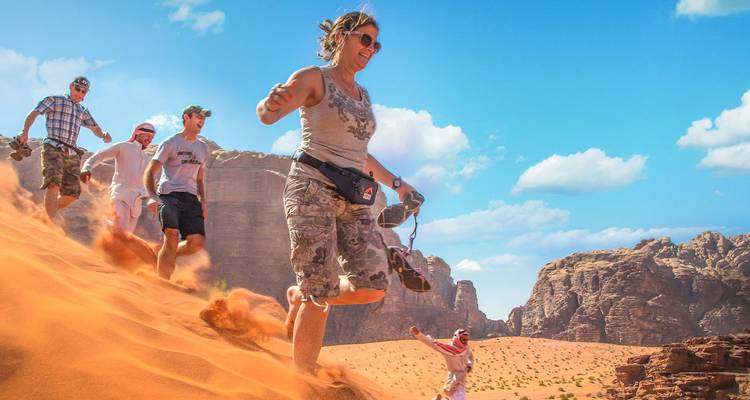 Des voyageurs riant à gorge déployée dégringolent rapidement une dune de sable orange abrupte dans le Wadi Rum sous un ciel lumineux.