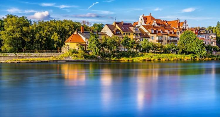 Les maisons colorées du village s'alignent le long d'une rive fluviale reflétée dans une eau lisse comme de la soie sous un ciel lumineux.