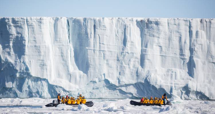 Two zodiacs of explorers navigate sea ice beside a towering vertical glacier wall.