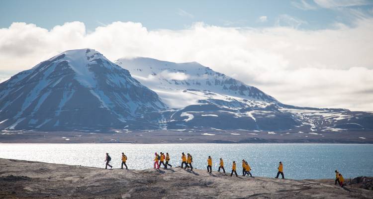 Hiking group in yellow jackets walks across rocky Arctic coast with snow-capped mountain backdrop.