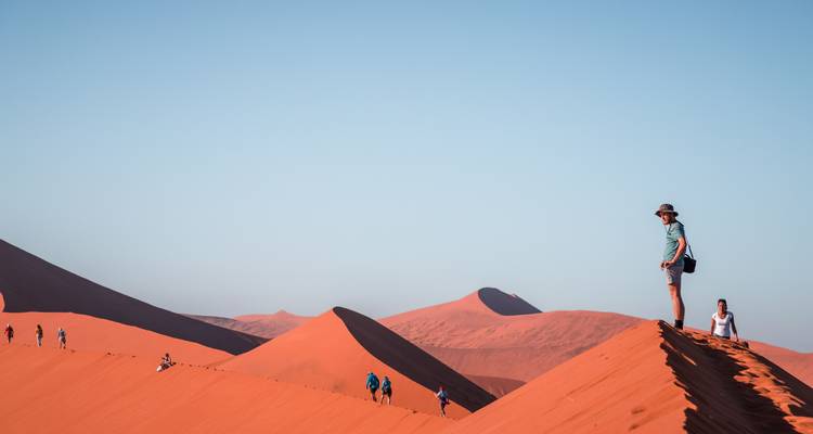 Wandelaars die een grote zandduinen afdalen onder een heldere hemel.