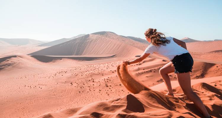 Vrouw zittend op een zandduin, zand gietend, met uitgestrekte duinen voor haar.