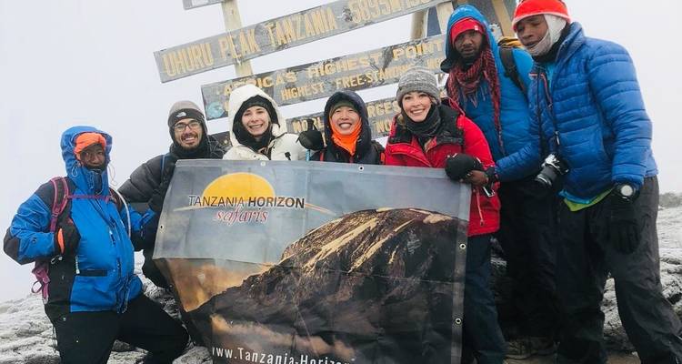 A group of climbers posing with a Mount Kilimanjaro sign and a banner.