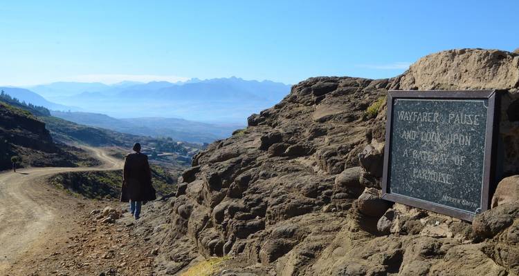 Personne debout sur un sentier rocheux avec un paysage de montagne pittoresque en arrière-plan et un panneau.