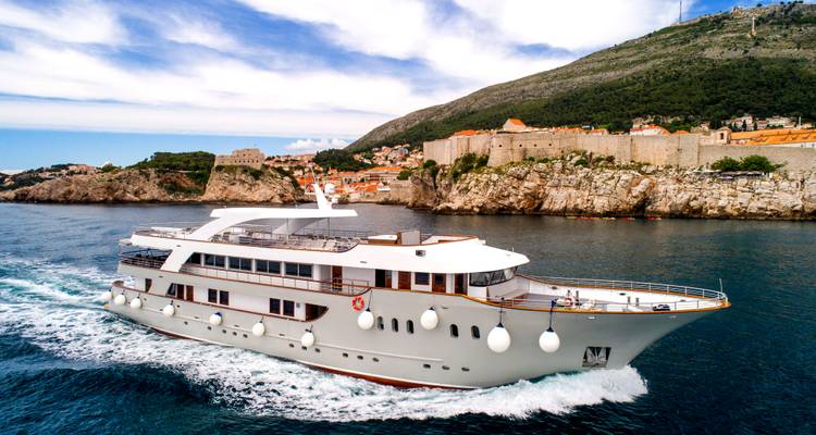 Un élégant yacht de croisière blanc file rapidement devant les anciens murs de pierre de Dubrovnik et sa ville sur les collines.