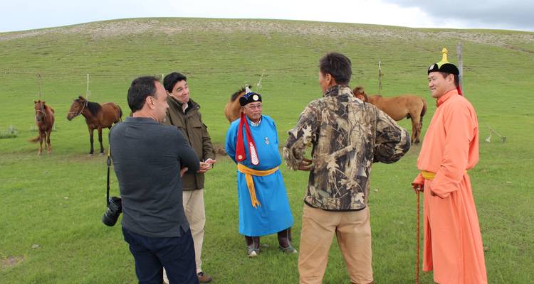 Grupo de personas con vestimenta tradicional mongola conversando en un área cubierta de hierba con caballos.