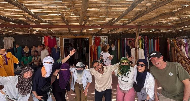 Un grupo de viajeros con pañuelos del desierto posa de forma divertida fuera de un puesto de mercado lleno de telas coloridas.
