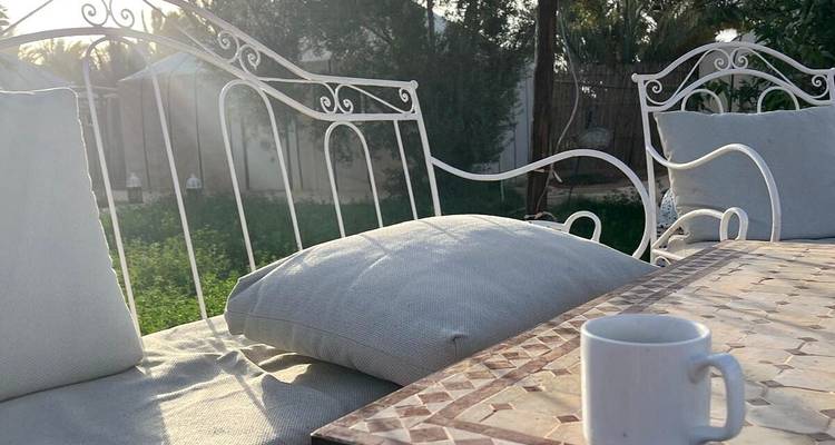 Escena de patio sombreado con banco de hierro forjado blanco, cojines y una taza de café matutino.