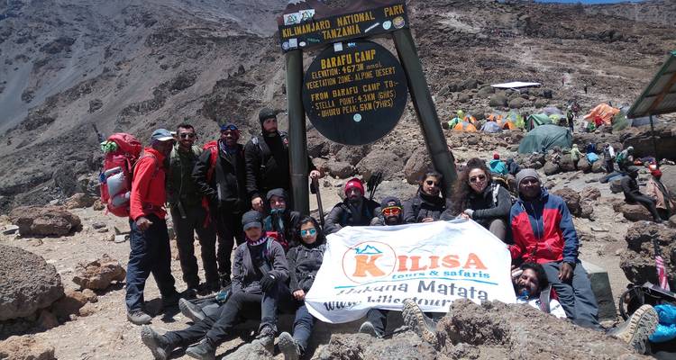 Un gran grupo de senderismo reunido en el letrero del Campamento Barafu en el Monte Kilimanjaro rodeado de terreno rocoso y tiendas de campaña.