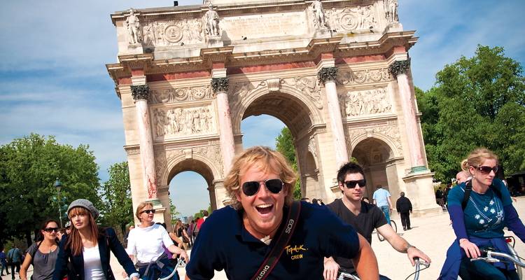 Cyclists ride past the Arc de Triomphe du Carrousel in Paris on a sunny day, led by a smiling man in the foreground