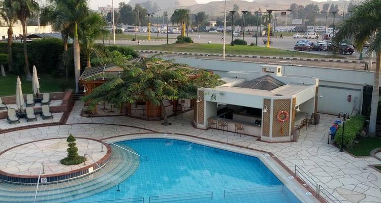 Hotel outdoor pool surrounded by loungers, palms and a snack bar with city traffic beyond the wall