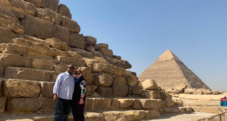 Smiling couple posing beside massive limestone blocks of a pyramid with another pyramid rising in the background under a blue sky