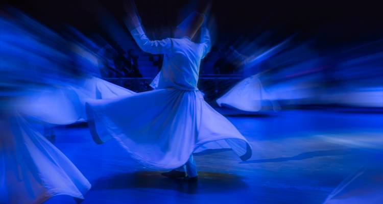 Derviche tourneur aux contours flous, vêtu de blanc, se produisant sous des projecteurs de scène bleus.