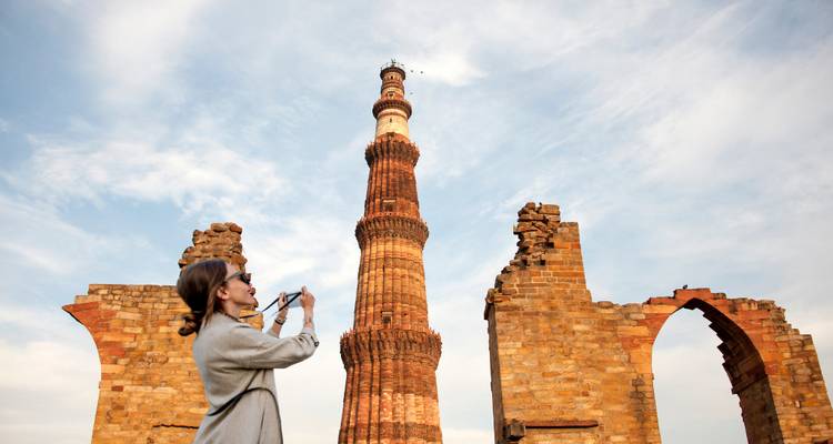Woman photographing towering Qutub Minar framed by ancient brick arches under blue sky
