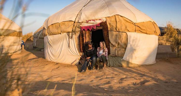 Twee reizigers die buiten een traditionele woestijnyurt bij zonsondergang met elkaar praten, overal zandduinen.