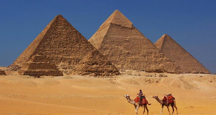 Trois pyramides de Giza avec deux chameaux et cavaliers traversant les sables dorés du désert sous un ciel bleu dégagé