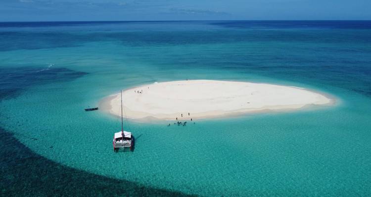 Vista aérea de una diminuta isla de arena blanca rodeada de agua turquesa vivida, con un catamarán anclado cerca y gente caminando en la arena.