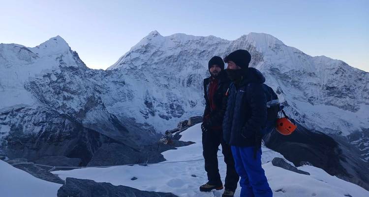 Zwei Kletterer stehen auf einem schneebedeckten Gipfel mit Bergen im Hintergrund.