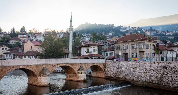 Un pont en arc de pierre et un minaret élancé s'élèvent au-dessus d'une rivière calme à Sarajevo au lever du soleil.