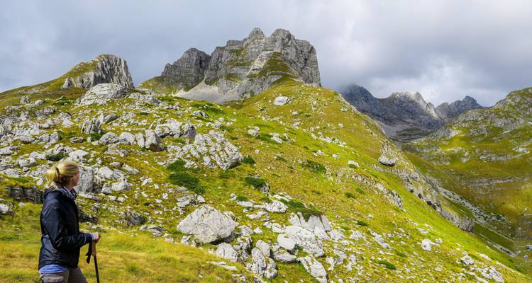Un randonneur observe les pics calcaires escarpés et les prairies vertes sous un ciel menaçant dans le Durmitor.