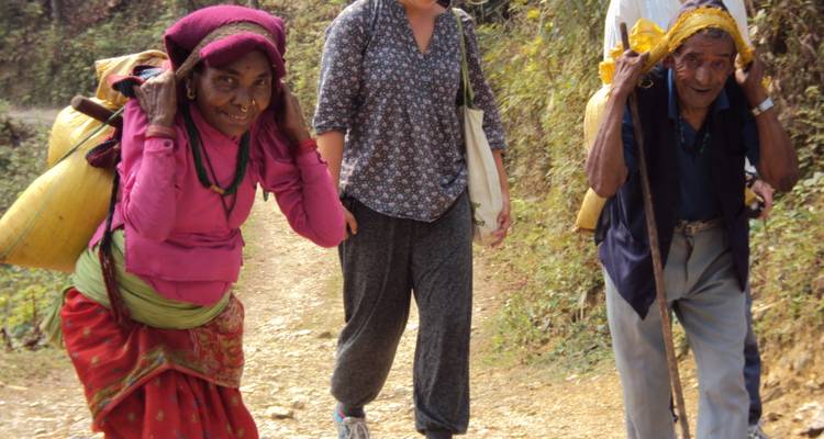 Dos ancianos del pueblo cargando sacos pesados pasan a un excursionista en un camino rural de montaña.