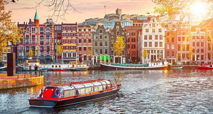 Un barco de crucero por canal con techo de cristal se desliza por un canal de Ámsterdam bordeado de edificios altos con frontones al atardecer.