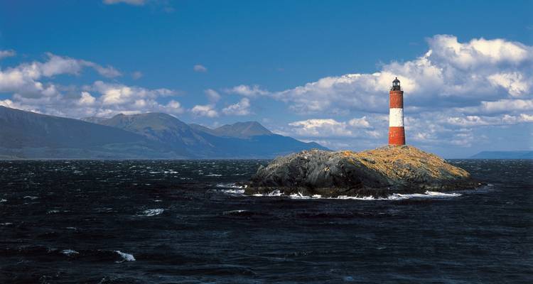 Faro en una isla rocosa con montañas al fondo.