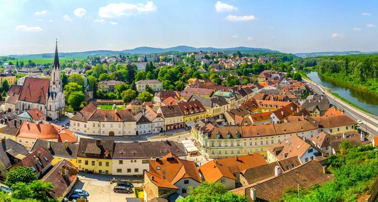 Riverside Austrian town with church spire and red-roofed houses set among rolling hills.