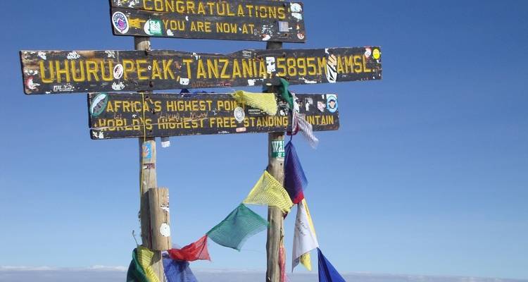 Cartel en el Pico Uhuru, Tanzania con banderas coloridas contra un cielo azul.