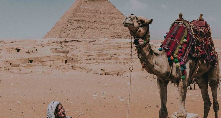Frau, die neben einem Kamel vor den Pyramiden sitzt.