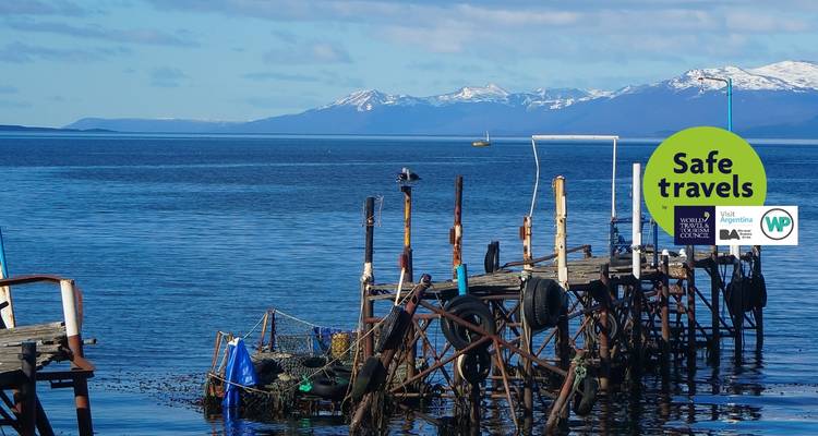 Ein rustikaler Steg mit einem "Gute Reise"-Aufkleber, der sich ins Wasser erstreckt mit Bergen im Hintergrund.