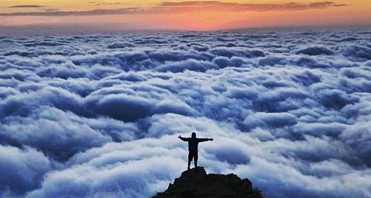Persoon op een bergtop omringd door wolken tijdens zonsondergang.
