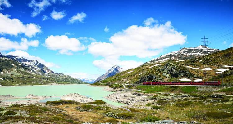 Un train rouge voyage à côté d'un réservoir alpin vert pâle entouré de pics couverts de neige.