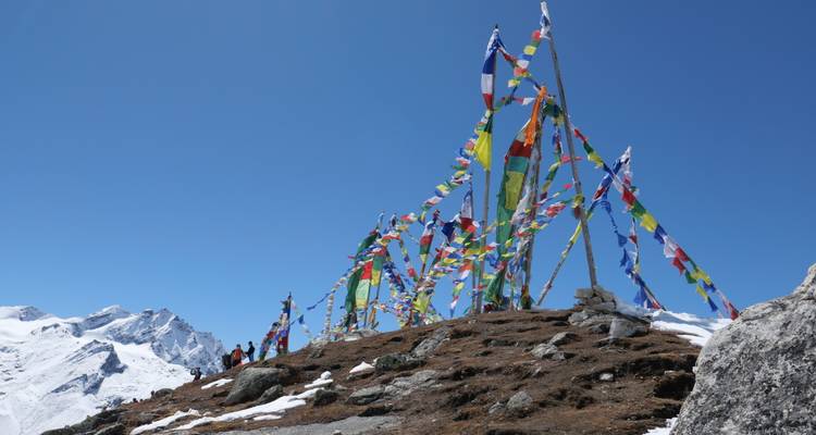 Gebetsflaggen flattern auf einem windumtosten Bergrücken, darunter Trekker und schneebedeckte Himalaya-Gipfel in der Ferne.