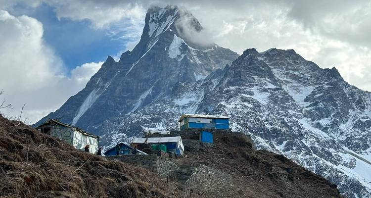 Small blue stone lodges cling to a rugged hillside beneath the dramatic snowy summit of Machhapuchhre shrouded in drifting cloud