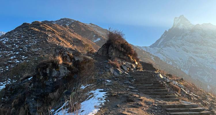 Stone steps of a mountain trail winding up a snow-speckled ridge with morning light on a distant Himalayan peak
