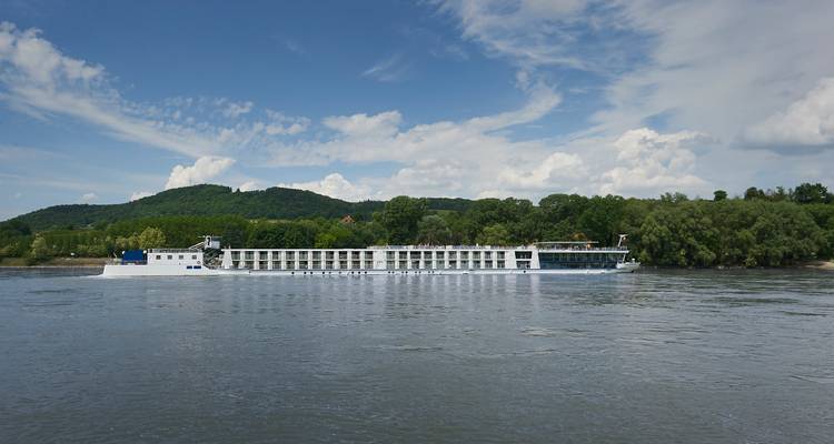 Long bateau de croisière naviguant sur une rivière avec une colline verdoyante en arrière-plan.