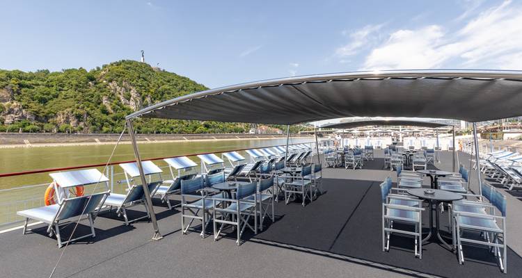 Spacious sundeck of a river cruise ship lined with lounge chairs under a canopy while cruising a calm river valley.